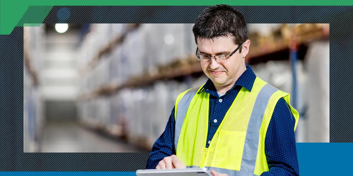 Man in a reflective vest using a smartphone in a warehouse aisle. Shelves filled with boxes blur into the background, conveying an industrial setting.