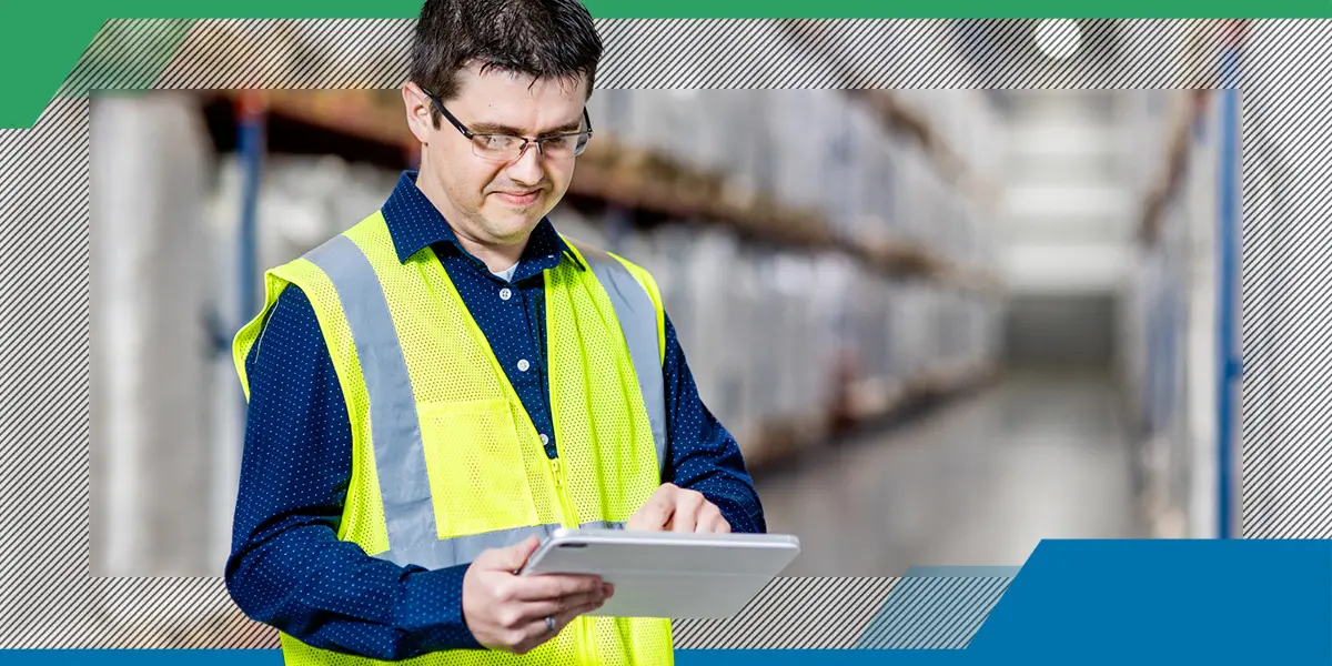 A man in a reflective vest holds a tablet, standing in a warehouse aisle. He appears focused, with shelves of inventory blurred in the background.