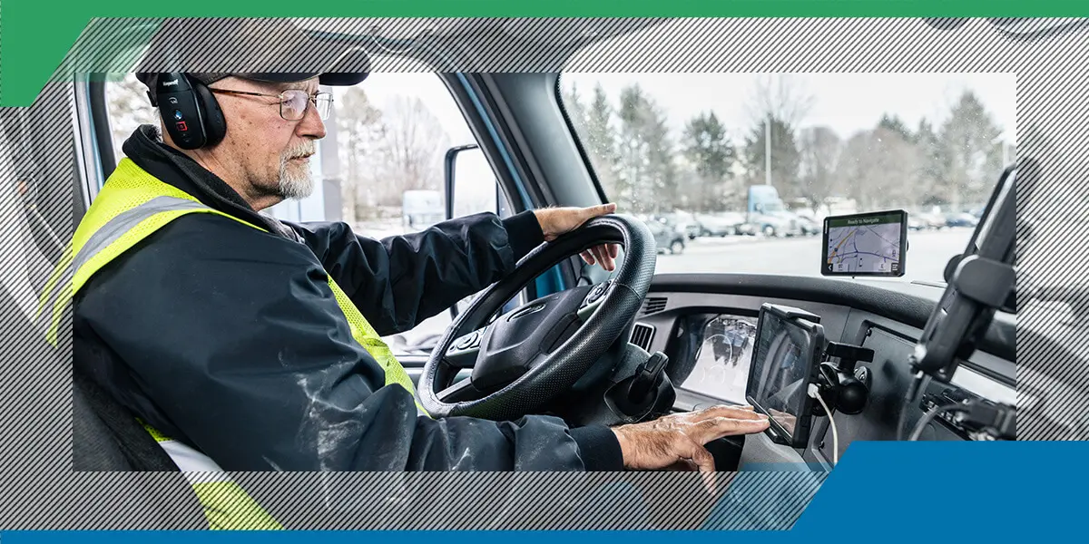 Driver in a high-visibility vest operates a truck, focusing on dashboard screens. The snowy scenery outside suggests winter driving conditions.